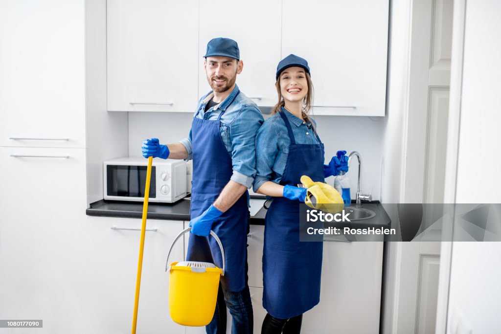 Portrait of a couple as a professional cleaners in uniform standing together with cleaning tools indoors